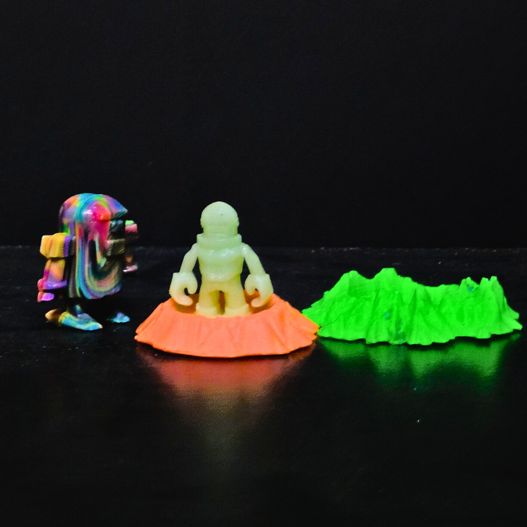 A swirly, multicolored water-heater style robot, a glow-in-the-dark spaceman, and two pieces of lunar terrain. They're all toys. It's hard to say from the photo how big they are, but I can say because I made them. The spaceman is about 50mm tall, and the robot about 60mm, and the lunar craters are roughly 100mm wide. 